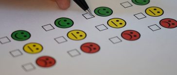 Main tenant un stylo pour cocher une case sur un papier d'enquête avec des icônes colorées de smiley et de visage triste indiquant différents niveaux de satisfaction au travail.