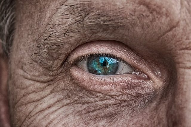 Gros plan sur l'œil d'une personne âgée à l'iris bleu et à la peau ridée, mettant en avant des produits adaptés au vieillissement cutané.