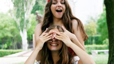 Deux jeunes femmes, l’une portant une casquette rouge et l’autre une chemise blanche, se livrent à une interaction ludique en plein air. L’une couvre les yeux de l’autre tout en souriant.