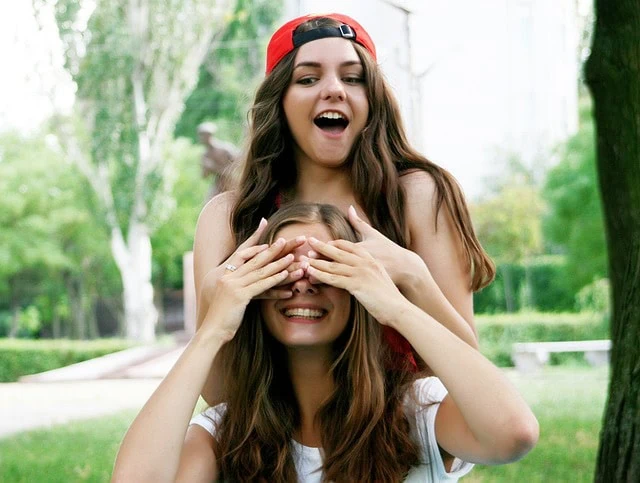 Deux jeunes femmes, l’une portant une casquette rouge et l’autre une chemise blanche, se livrent à une interaction ludique en plein air. L’une couvre les yeux de l’autre tout en souriant.