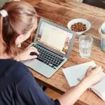 Une femme, mettant en valeur la qualité essentielle de l'empathie, travaille comme rédactrice sur son ordinateur portable à une table en bois, entourée d'un cahier, d'un verre d'eau, d'amandes et d'une corbeille de fruits avec des ananas et des oranges.