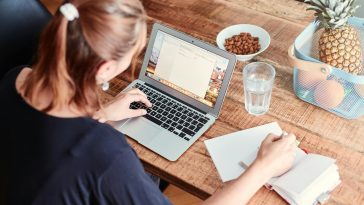 Une femme, mettant en valeur la qualité essentielle de l'empathie, travaille comme rédactrice sur son ordinateur portable à une table en bois, entourée d'un cahier, d'un verre d'eau, d'amandes et d'une corbeille de fruits avec des ananas et des oranges.