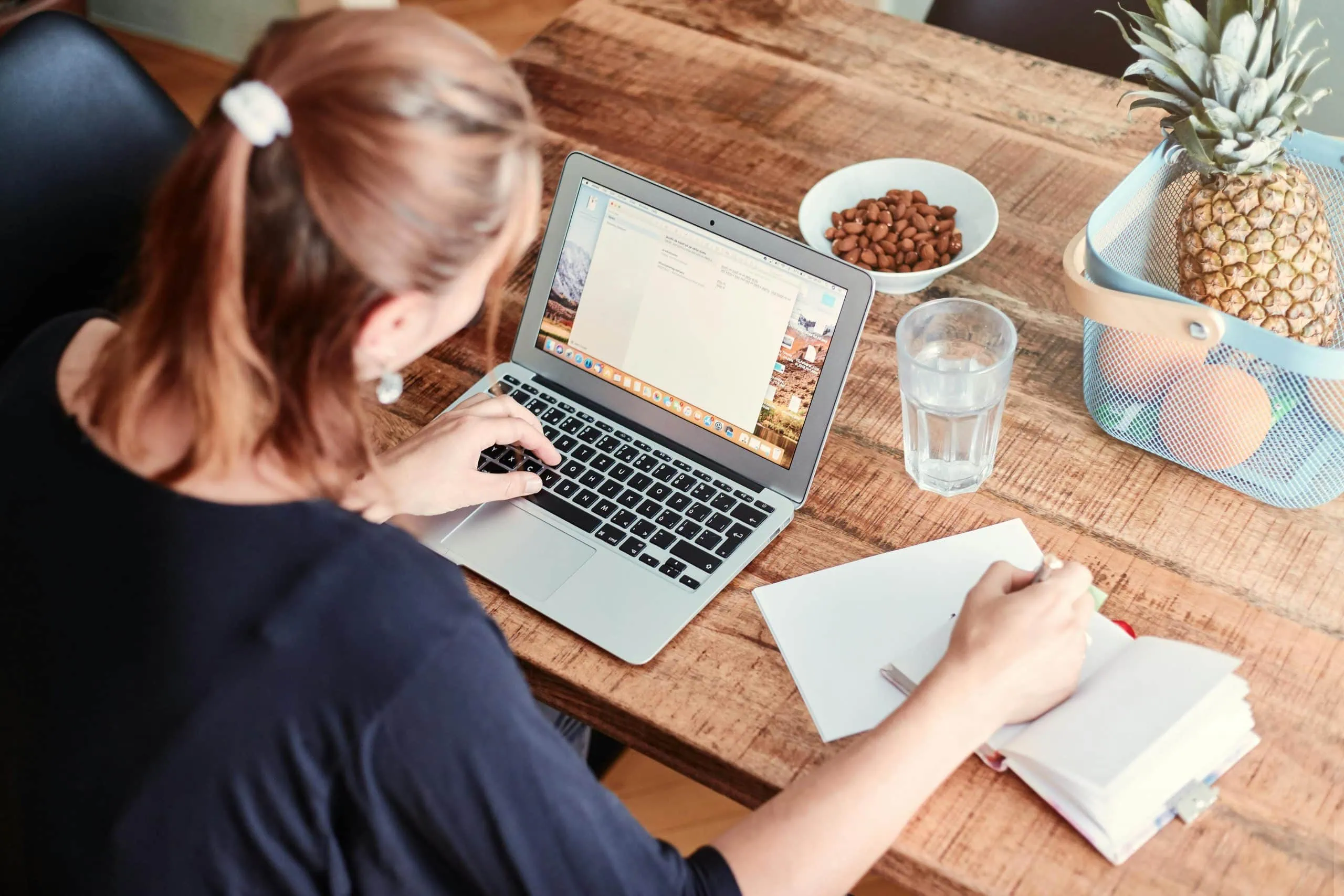 Une femme, mettant en valeur la qualité essentielle de l'empathie, travaille comme rédactrice sur son ordinateur portable à une table en bois, entourée d'un cahier, d'un verre d'eau, d'amandes et d'une corbeille de fruits avec des ananas et des oranges.