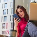 Une personne assise près d'une fenêtre avec un smartphone et un ordinateur portable, portant un haut rose. Une tasse de café à emporter est devant.