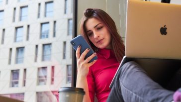 Une personne assise près d'une fenêtre avec un smartphone et un ordinateur portable, portant un haut rose. Une tasse de café à emporter est devant.