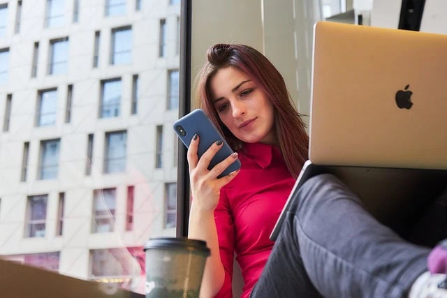 Une personne assise près d'une fenêtre avec un smartphone et un ordinateur portable, portant un haut rose. Une tasse de café à emporter est devant.